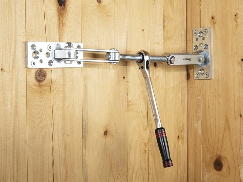 The Rothoblaas Geko Panel Puller, by Rothoblaas, is a metal hinge mechanism with handle mounted on wooden walls for precise assembly—ideal for adjusting or locking solid timber panels.