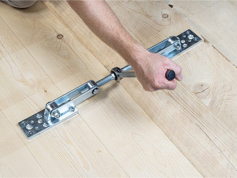 A person uses the Rothoblaas Geko Panel Puller, a floor clamp tool by Rothoblaas, to pull wooden floorboards tightly together during installation on a light-colored wood floor.