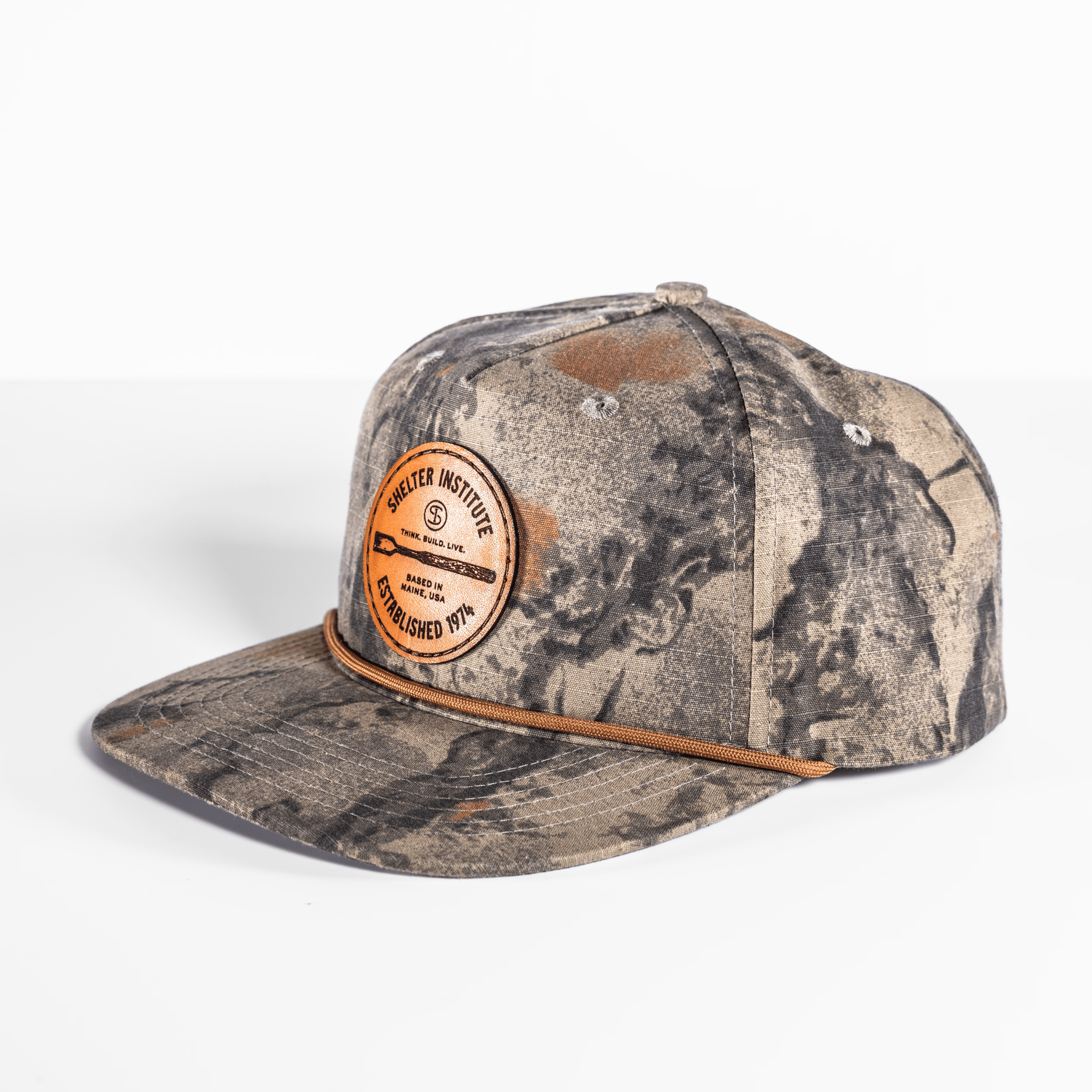 The Shelter Institute Camo Hat features a leather patch with text, a flat peak, and an adjustable back for comfort.