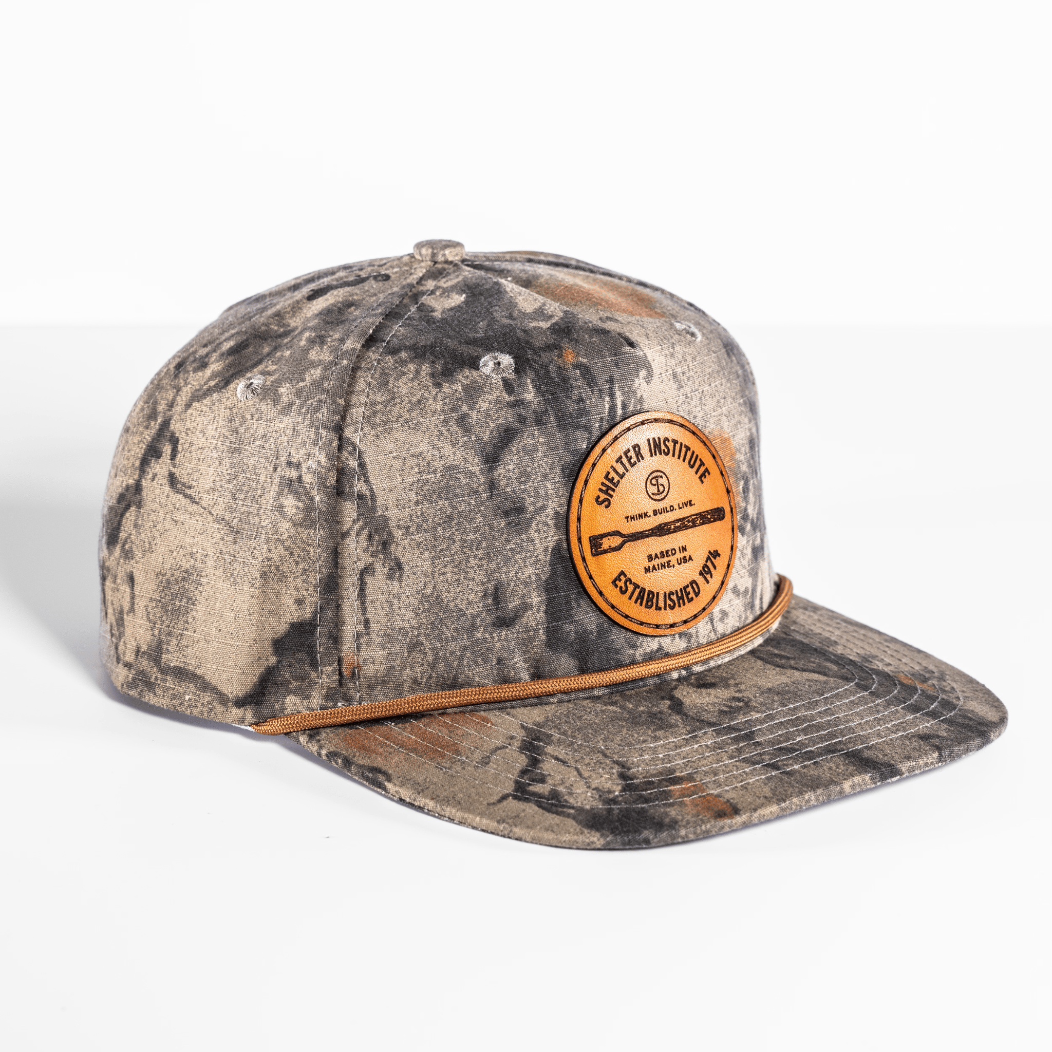 The Shelter Institute Camo Hat is a snapback featuring camouflage, a round orange "Shelter Institute, Established 1974" patch, and a rugged brown cord above the brim.