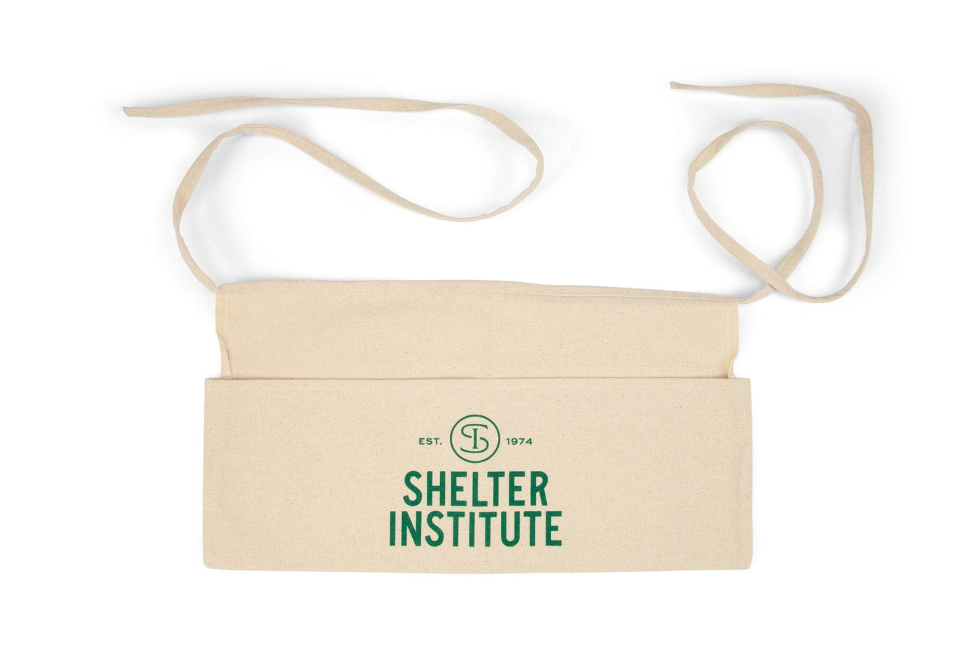 The Shelter Institute Cotton Apron is a natural cotton waist apron with long ties, a front pocket, and green Shelter Institute EST. 1974 text with circular logo—combining practicality and style from the Shelter Institute brand.