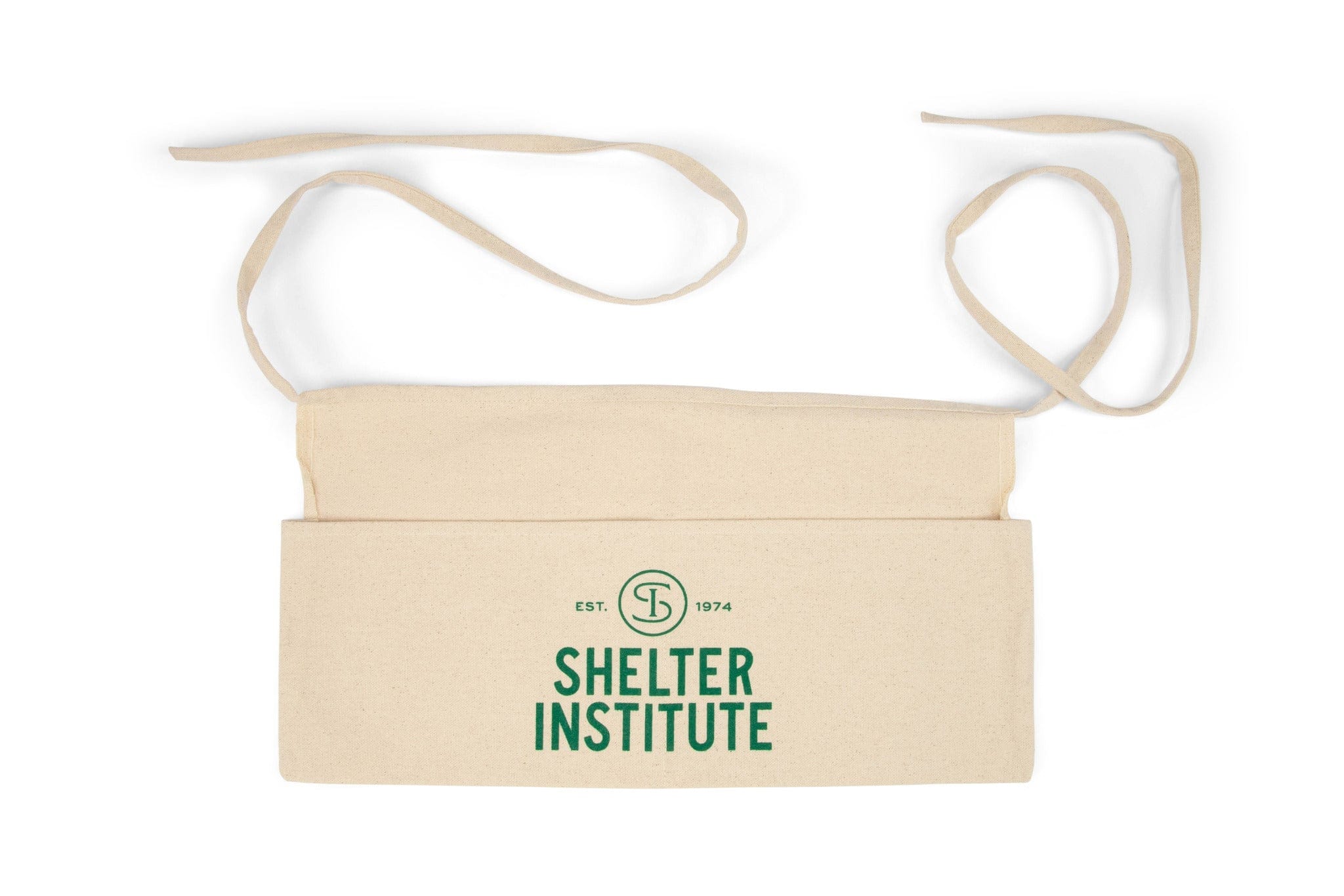 The Shelter Institute Cotton Apron is a natural cotton waist apron with long ties, a front pocket, and green Shelter Institute EST. 1974 text with circular logo—combining practicality and style from the Shelter Institute brand.