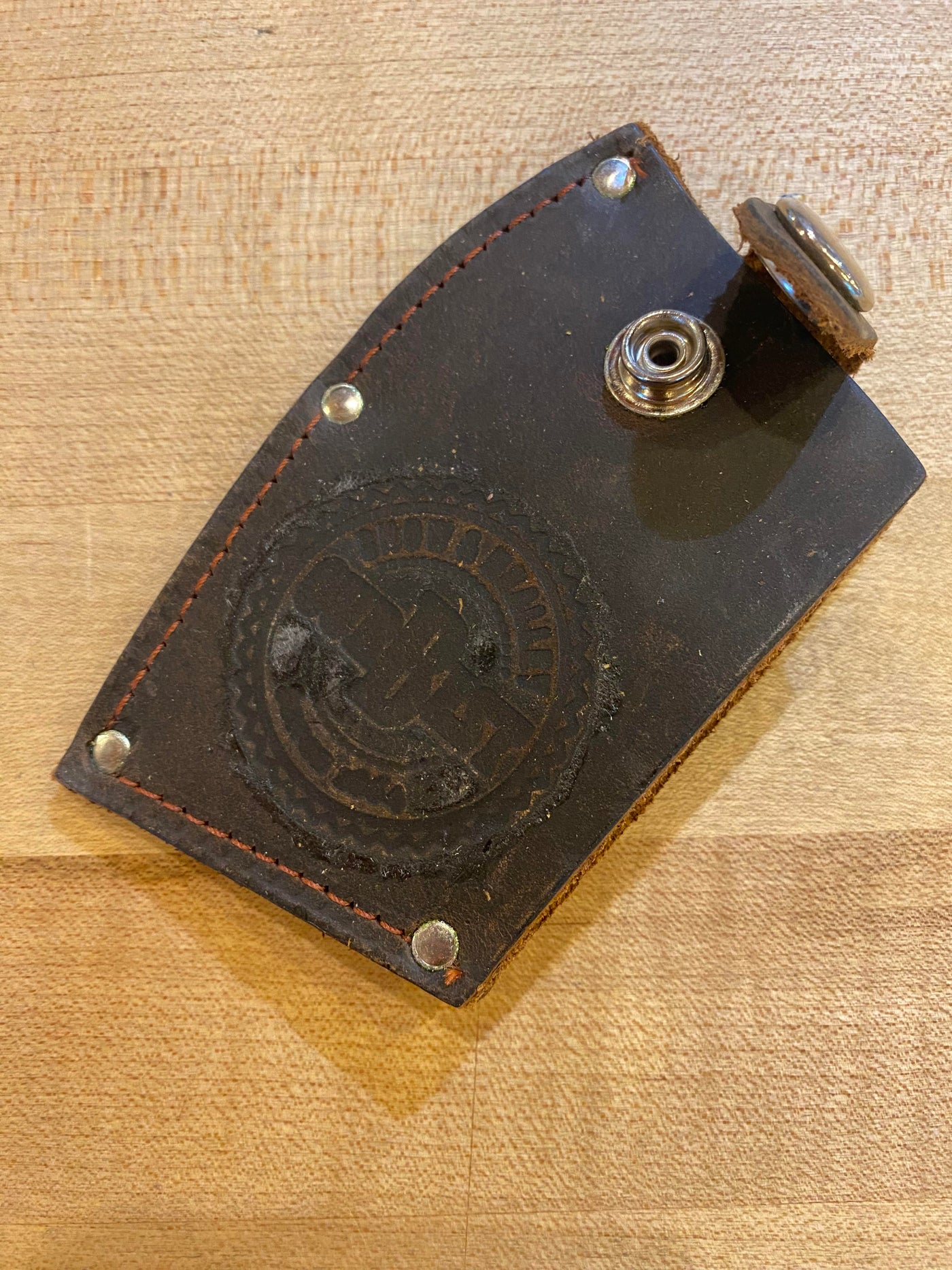 A brown leather key holder with a snap button closure, featuring a stamped emblem and several small rivets, is lying on a wooden surface next to the Snow & Nealley Pen Bay Kindling Axe by SNO, which boasts an American hickory handle.