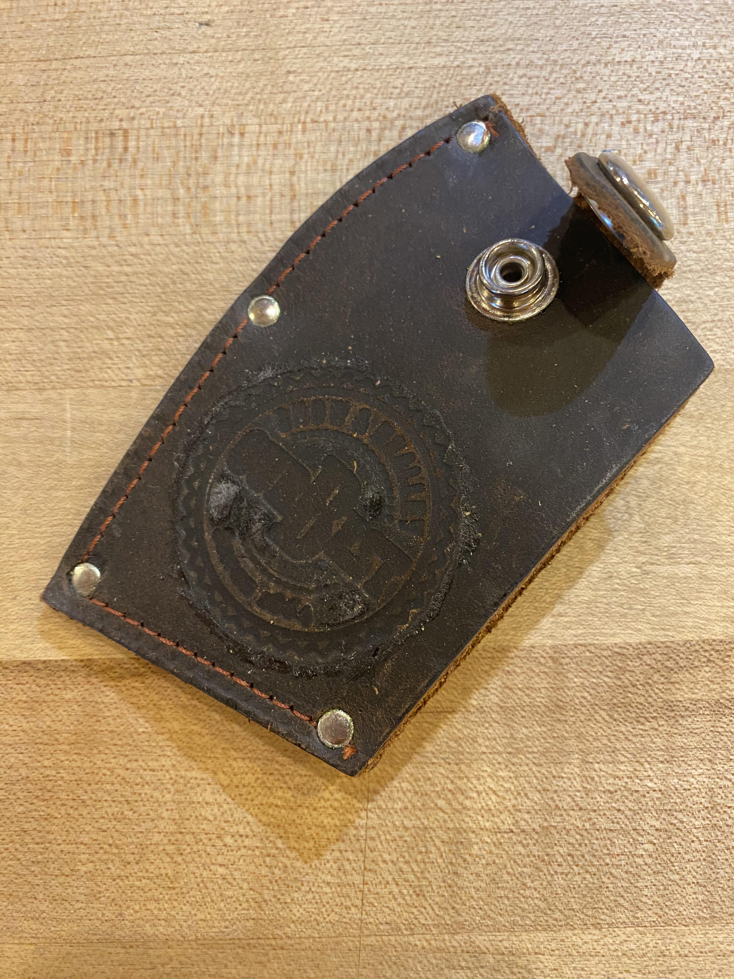 A brown leather key holder with a snap button closure, featuring a stamped emblem and several small rivets, is lying on a wooden surface next to the Snow & Nealley Pen Bay Kindling Axe by SNO, which boasts an American hickory handle.