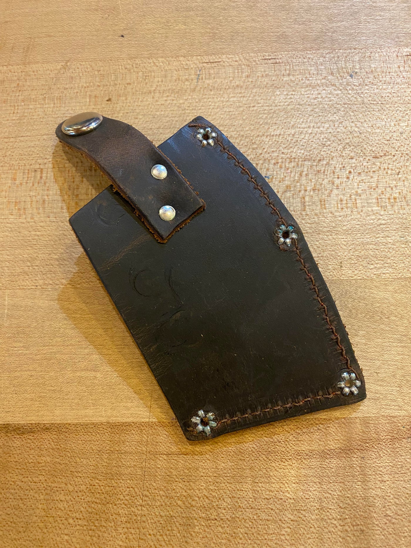 A black leather sheath with decorative stitching and four metal studs lies on a wooden surface. A strap with a metal ring is attached at the top, perfectly complementing the Snow & Nealley Pen Bay Kindling Axe's carbon steel axe head and American hickory handle from SNO.