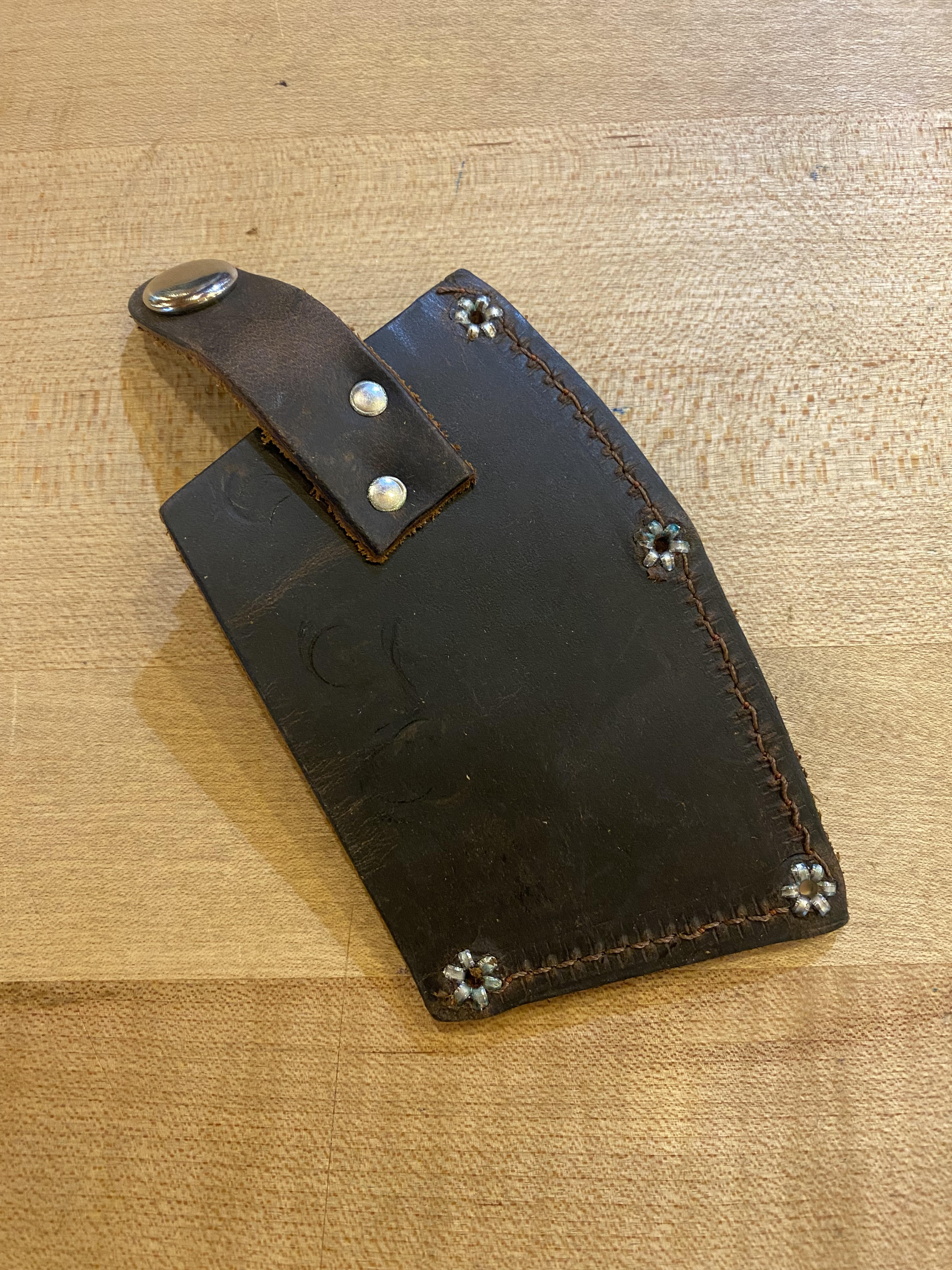 A black leather sheath with decorative stitching and four metal studs lies on a wooden surface. A strap with a metal ring is attached at the top, perfectly complementing the Snow & Nealley Pen Bay Kindling Axe's carbon steel axe head and American hickory handle from SNO.