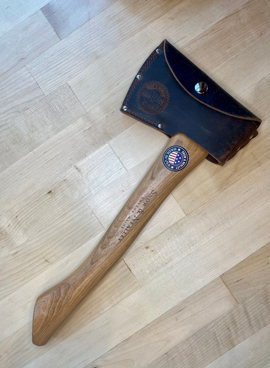 The SNO Campers Belt Axe, featuring a lightweight American hickory handle and leather sheath, rests on light wood. The handle shows Southampton and an American flag sticker—perfect for hiking and camping trips.