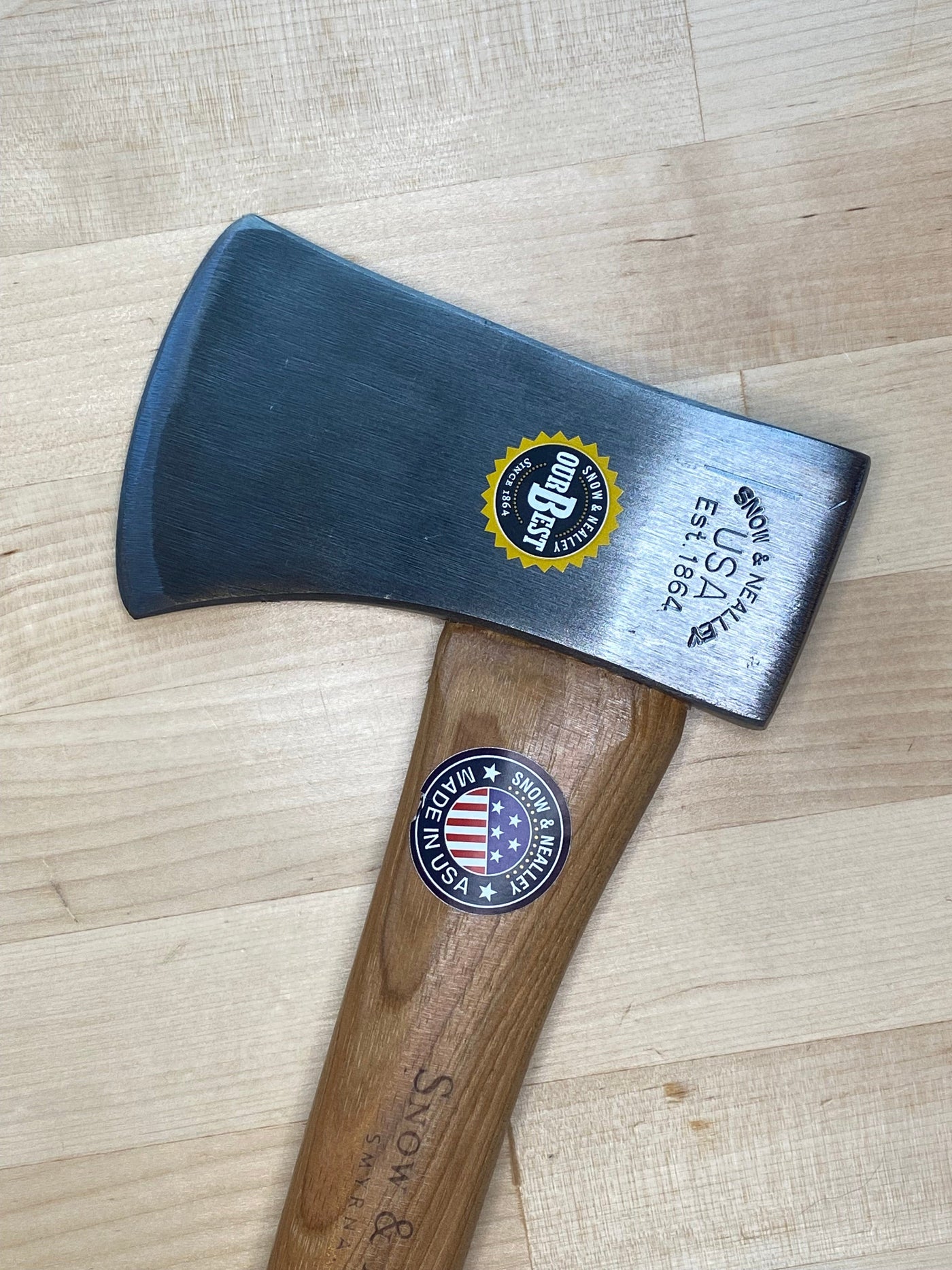 A close-up of SNOs Campers Belt Axe with a steel blade and American hickory handle rests on light wood. The blade shows Council Tool Co. USA and a 100 Best sticker, while the handle has a Made in USA sticker—perfect for hiking or camping.