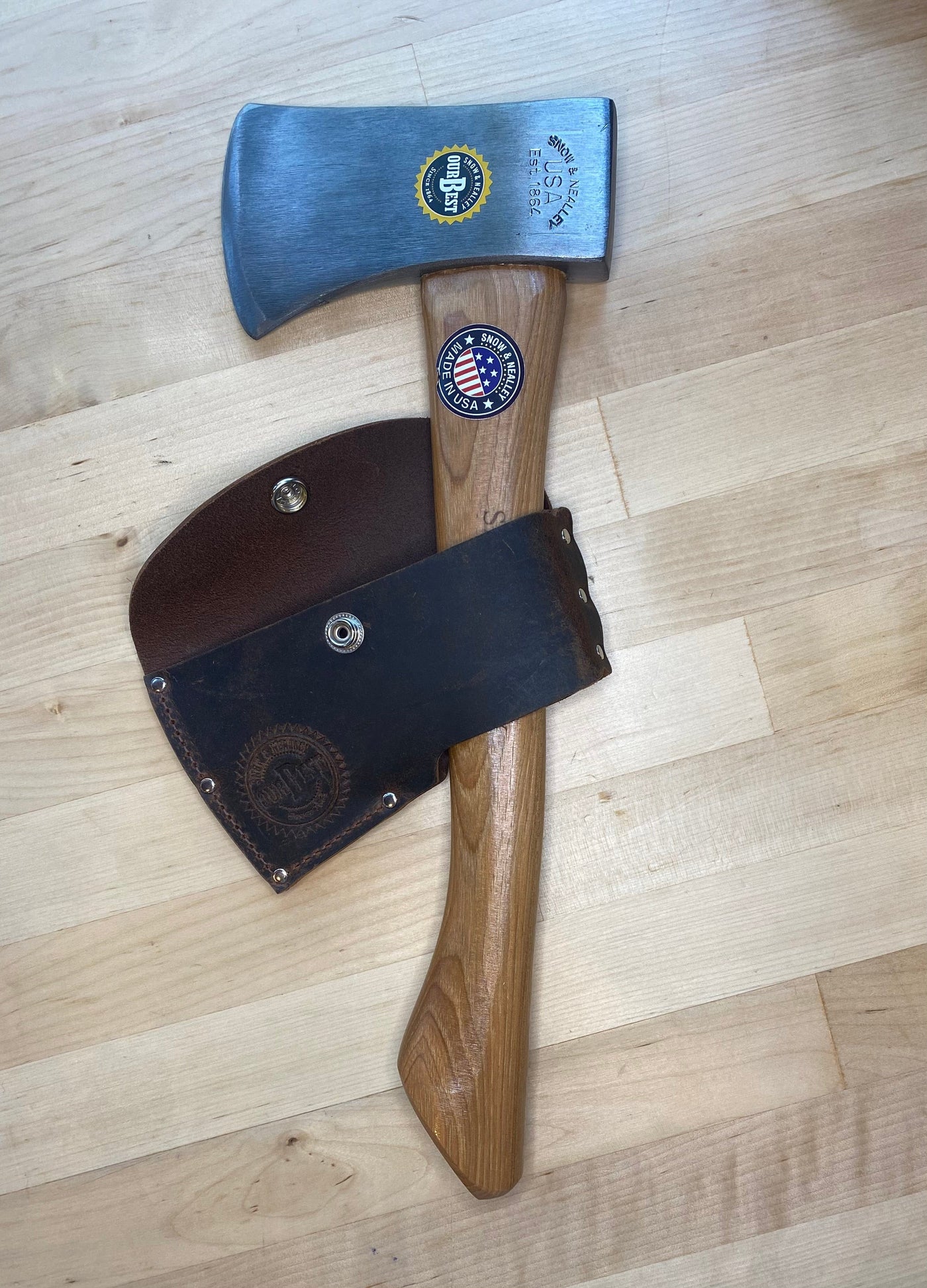 The SNO Campers Belt Axe, with an American hickory handle and steel blade adorned with stickers and engravings, rests on a light wooden surface. Its leather blade cover attaches to the handle—ideal for hiking or camping.
