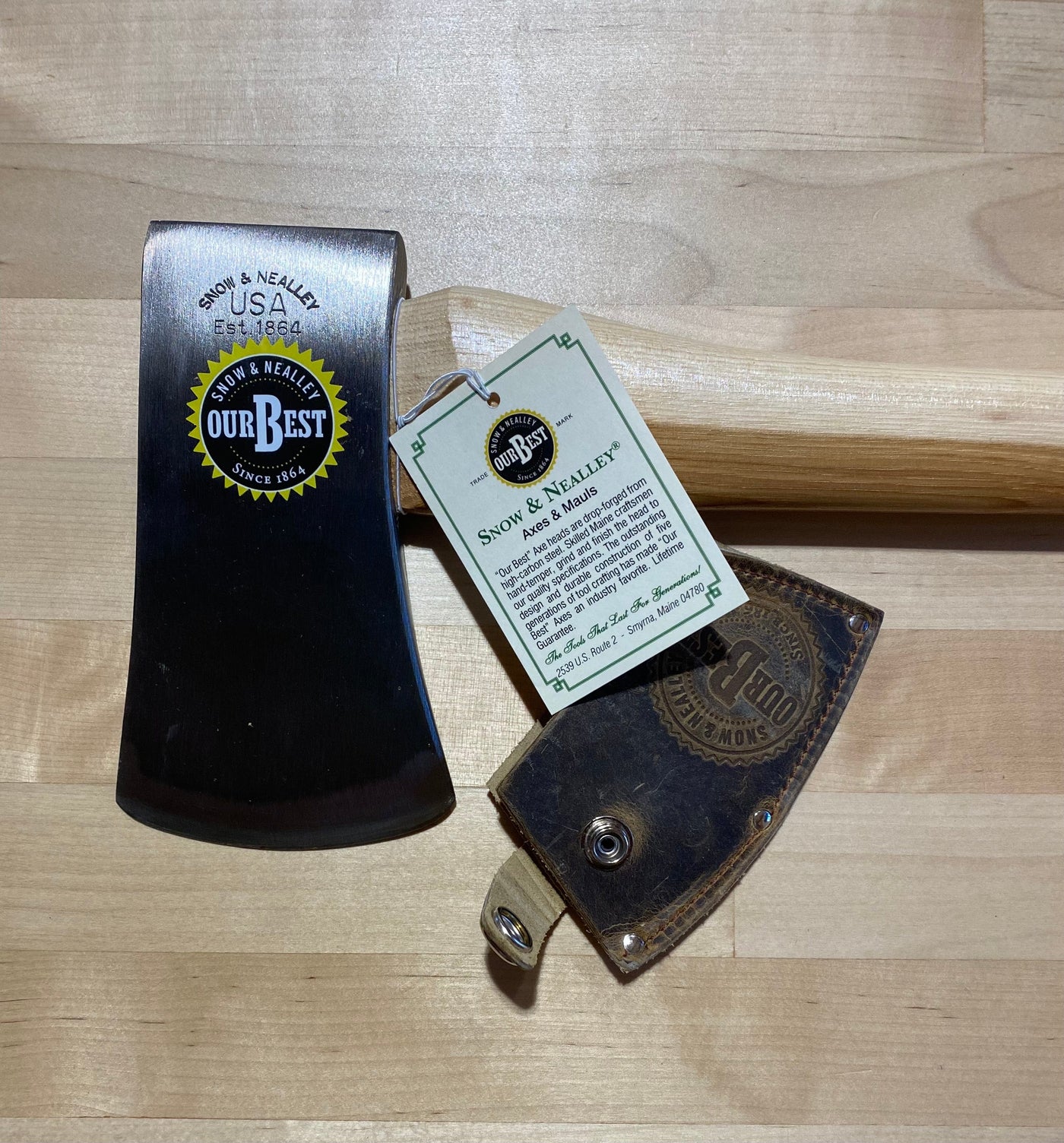 The SNO Snow & Neally Axe 2-1/4-Lb features a high-carbon steel head, American hickory handle, and leather sheath. Displayed on wood with an info tag attached.