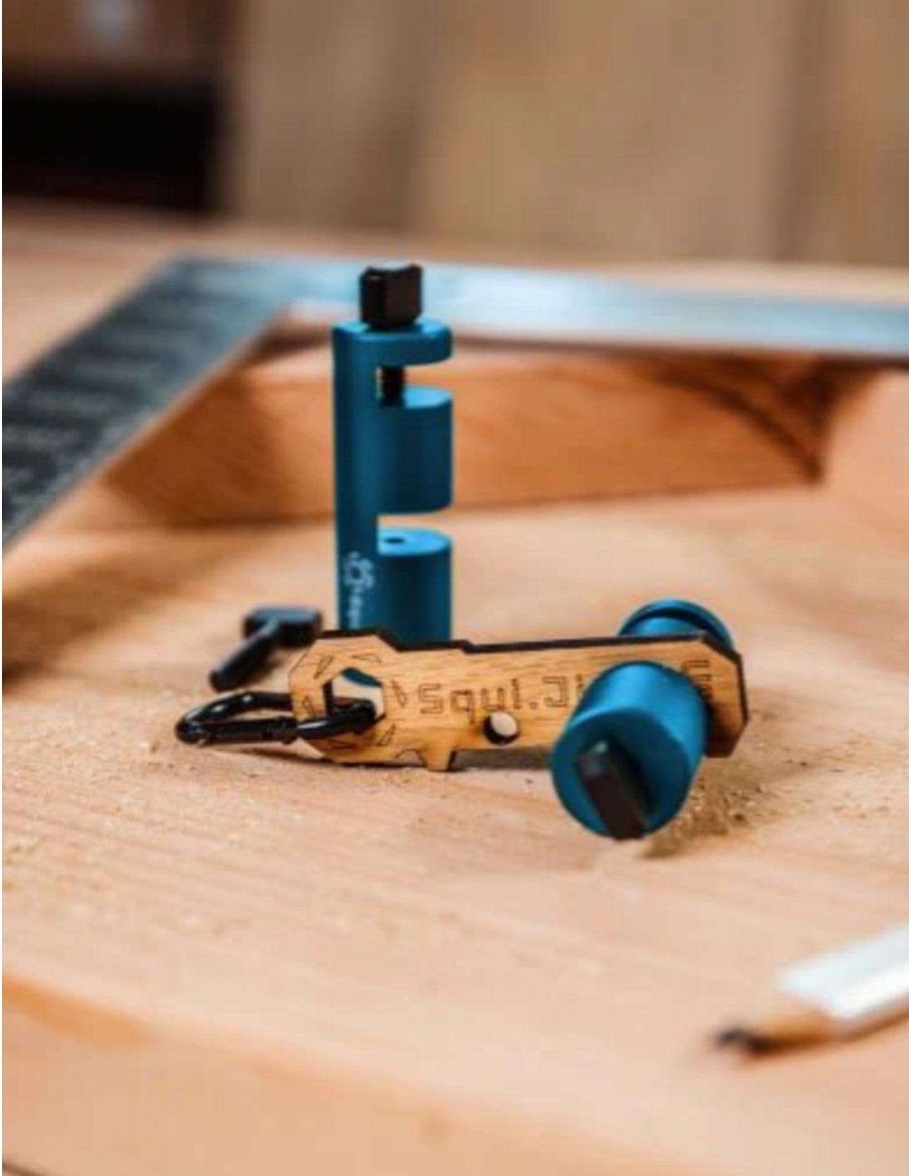 A close-up of Squi.Jig Framing Stair Gauge metal tools by Squi.Jig, shown with a wooden engraved keychain on sawdust, a ruler and pencil blurred in back—perfect for precision stair layout projects.