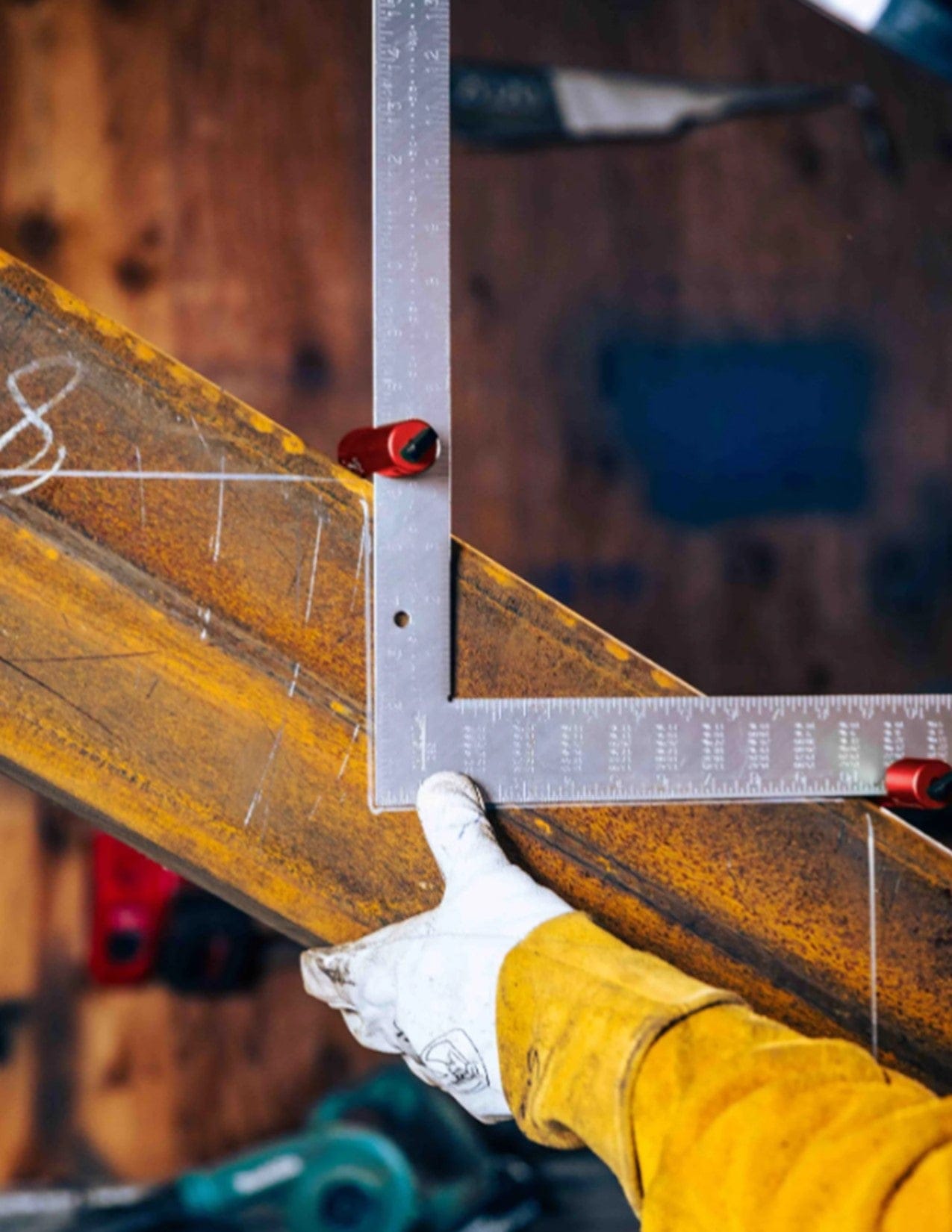 A person in a yellow jacket uses the Squi.Jig Framing Stair Gauge by Squi.Jig to mark an angled steel beam in a workshop with tools and wooden surfaces blurred in the background.