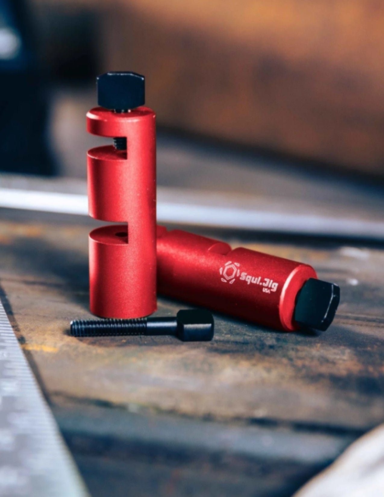 Two Squi.Jig Framing Stair Gauges by Squi.Jig with hexagonal ends and slots rest on a workbench beside a black screw and metal ruler, ideal for precise stair or rafter layout. The background is blurred.