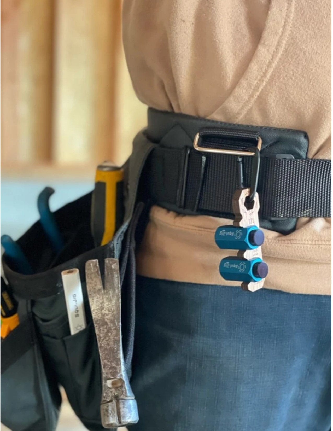 A close-up shows a person wearing a tool belt holding various tools, including a hammer, pliers, and Squi.Jig Framing Stair Guage by Squi.Jig. Two blue magnetic tool holders are attached to the belt; the background is blurred.
