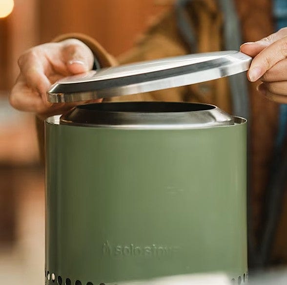 A person lifts the metal lid off a green SSO Mesa XL tabletop fire pit, ready to use it. The softly blurred background highlights their hands and the stove—ideal for pairing with the SSO Mesa XL Accessory Pack.