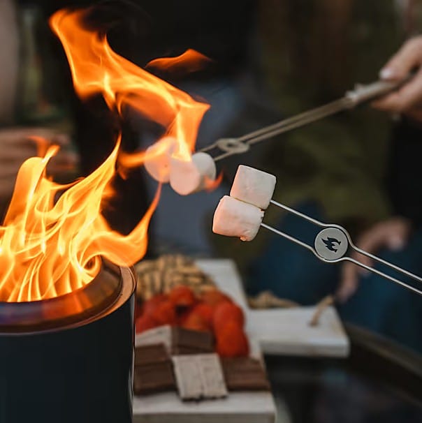 Two people roast marshmallows over the XL Mesa tabletop fire pit. An SSO Mesa XL Accessory Pack tray with chocolate, graham crackers, and strawberries is in the background—ideal for s’mores.