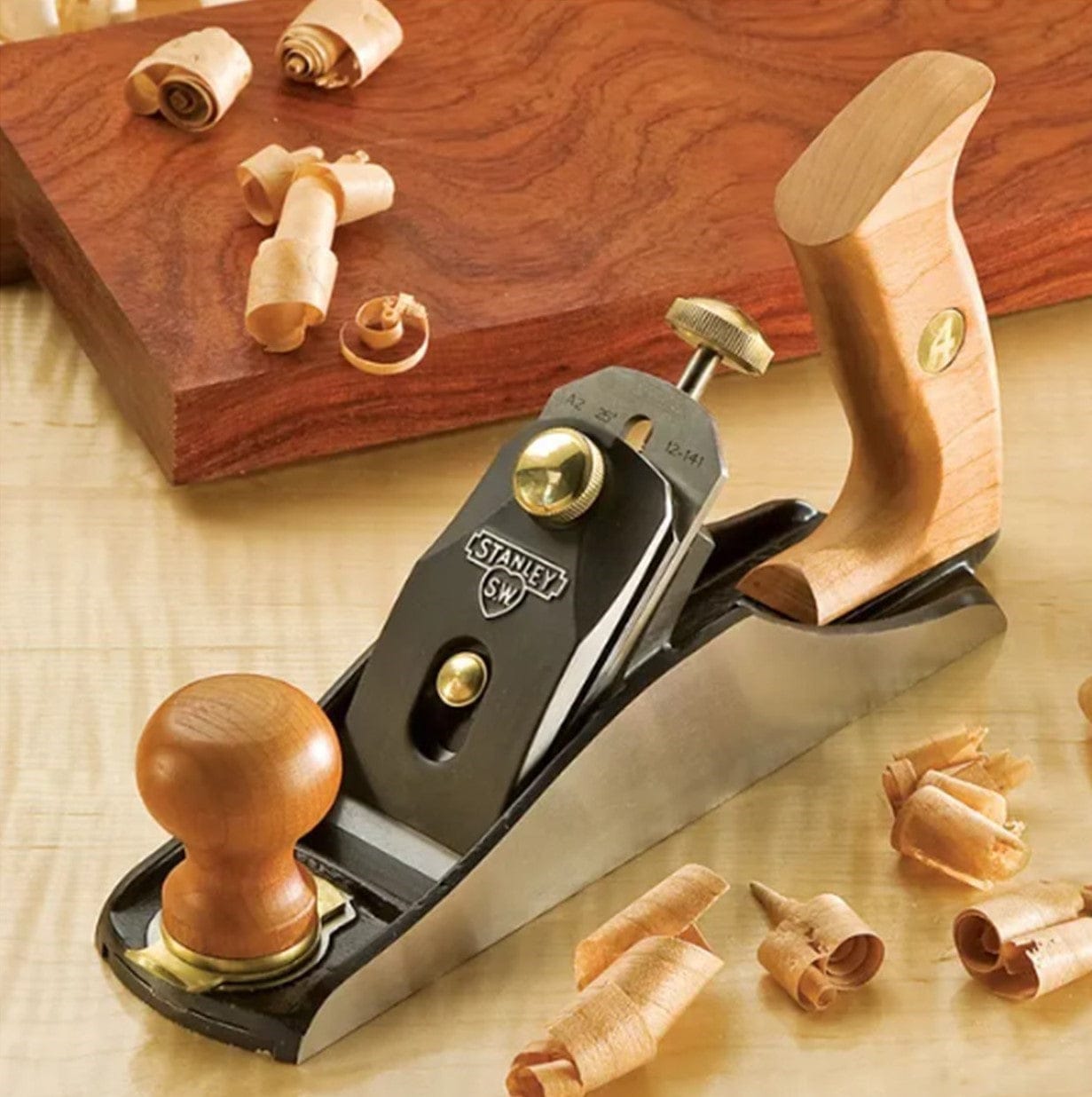 A STN No. 4 SweetHeart™ Smoothing Bench Plane with wooden handles sits on wood shavings and a finished board, featuring an adjustable throat plate for precise woodworking.