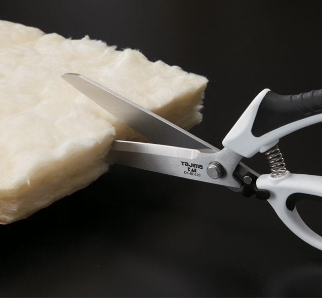 A pair of TAJ Tajima VARIX Single Ring Shears is cutting through a thick, fluffy sheet of light-colored material on a black surface.