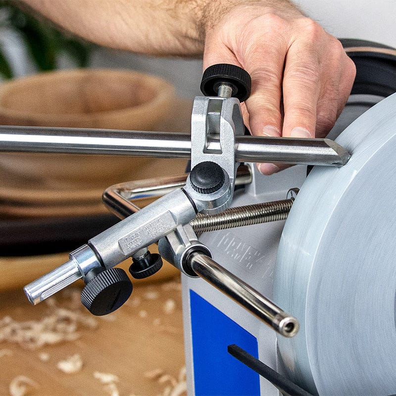 A person adjusts the TOR Tormek Gouge Jig’s metal guide while a woodturning tool rests against a spinning sharpening stone, with wood shavings and wooden bowls on a nearby workbench.