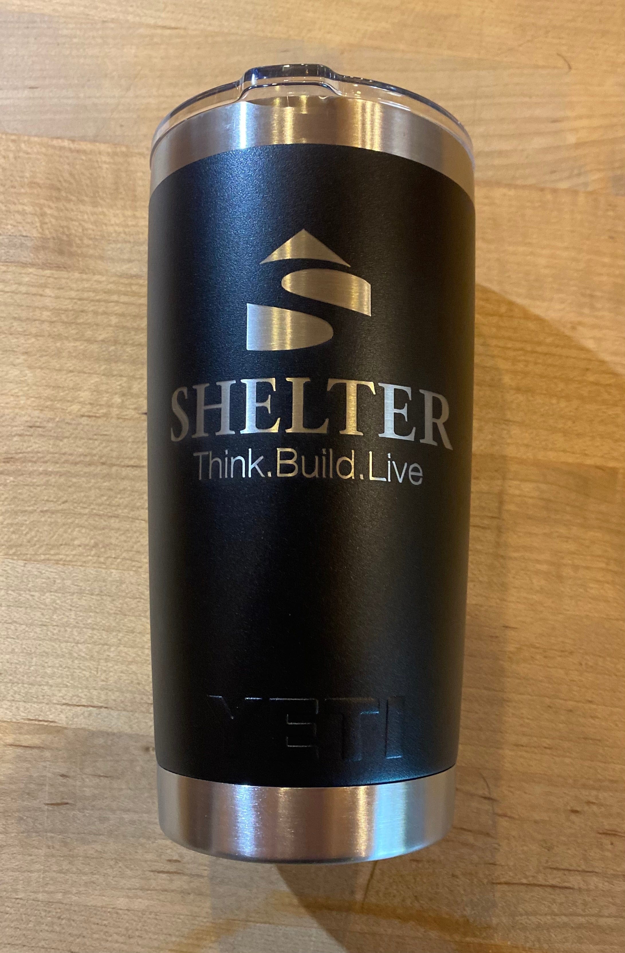 The Shelter Yeti Rambler Tumbler is a durable insulated black tumbler featuring a roof logo and the phrase "Shelter Think. Build. Live." Made from stainless steel with double-wall vacuum insulation, it has "YETI" engraved at the bottom and rests elegantly on a wooden surface.