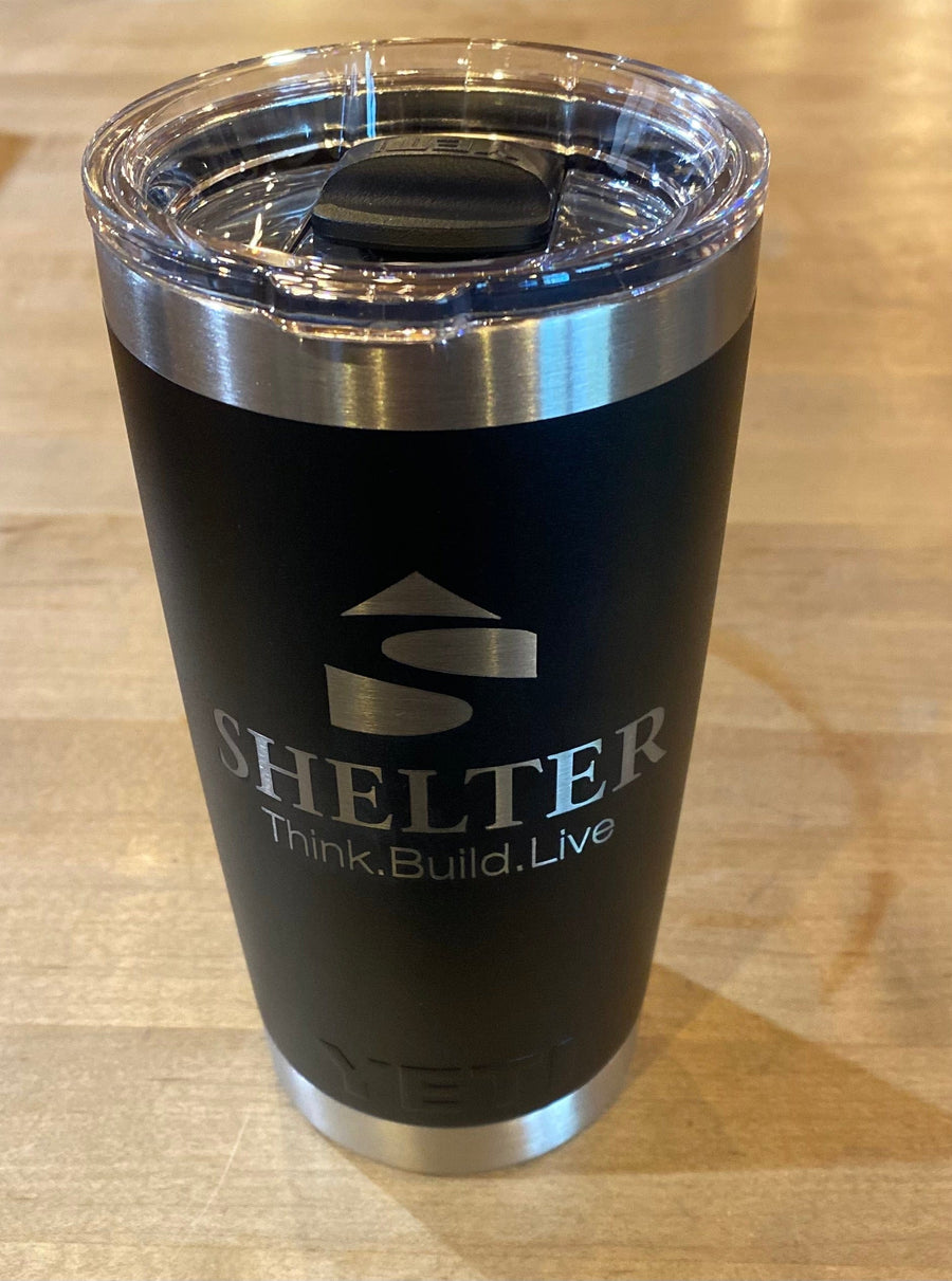 Black and silver Shelter Yeti Rambler Tumbler, featuring durable stainless steel and double-wall vacuum insulation, with the "Shelter Think. Build. Live." logo on a wooden surface.
