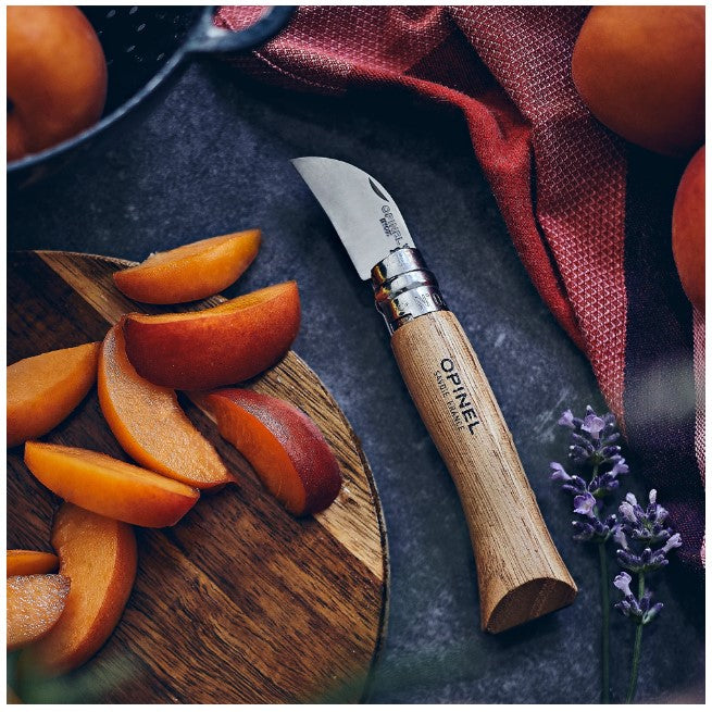 An Opinel No.07 Folding Chestnut and Garlic Knife with a wooden handle and stainless steel blade rests beside sliced plums on a round wooden board. Nearby are whole plums, a red cloth, and sprigs of lavender on a dark surface.