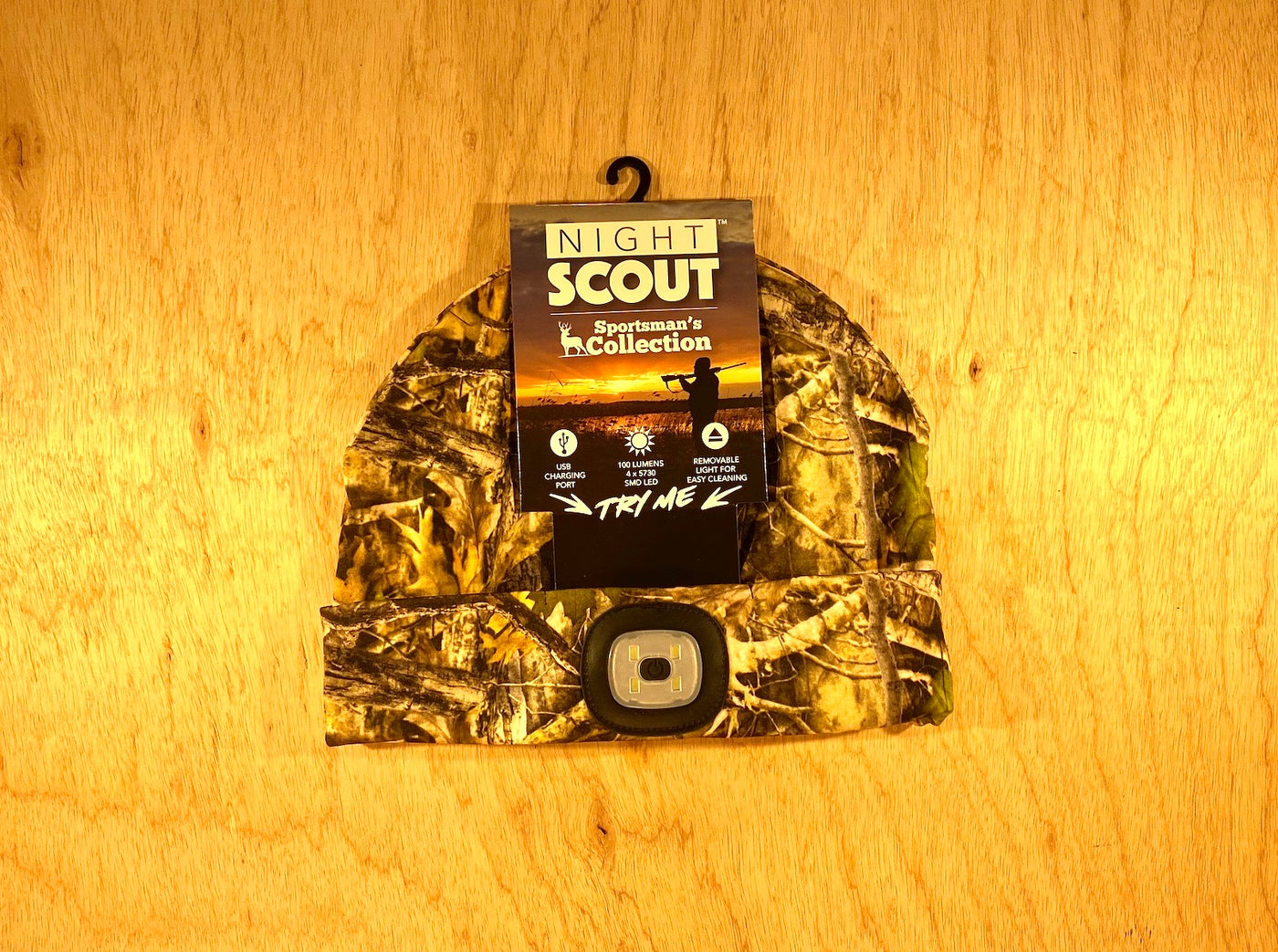 The Shelter Institute Night Scout Camo Hunting Headlamp Beanie is shown with its built-in LED light against a wooden background. The label shows the Night Scout Sportsmans Collection and a sunset silhouette.