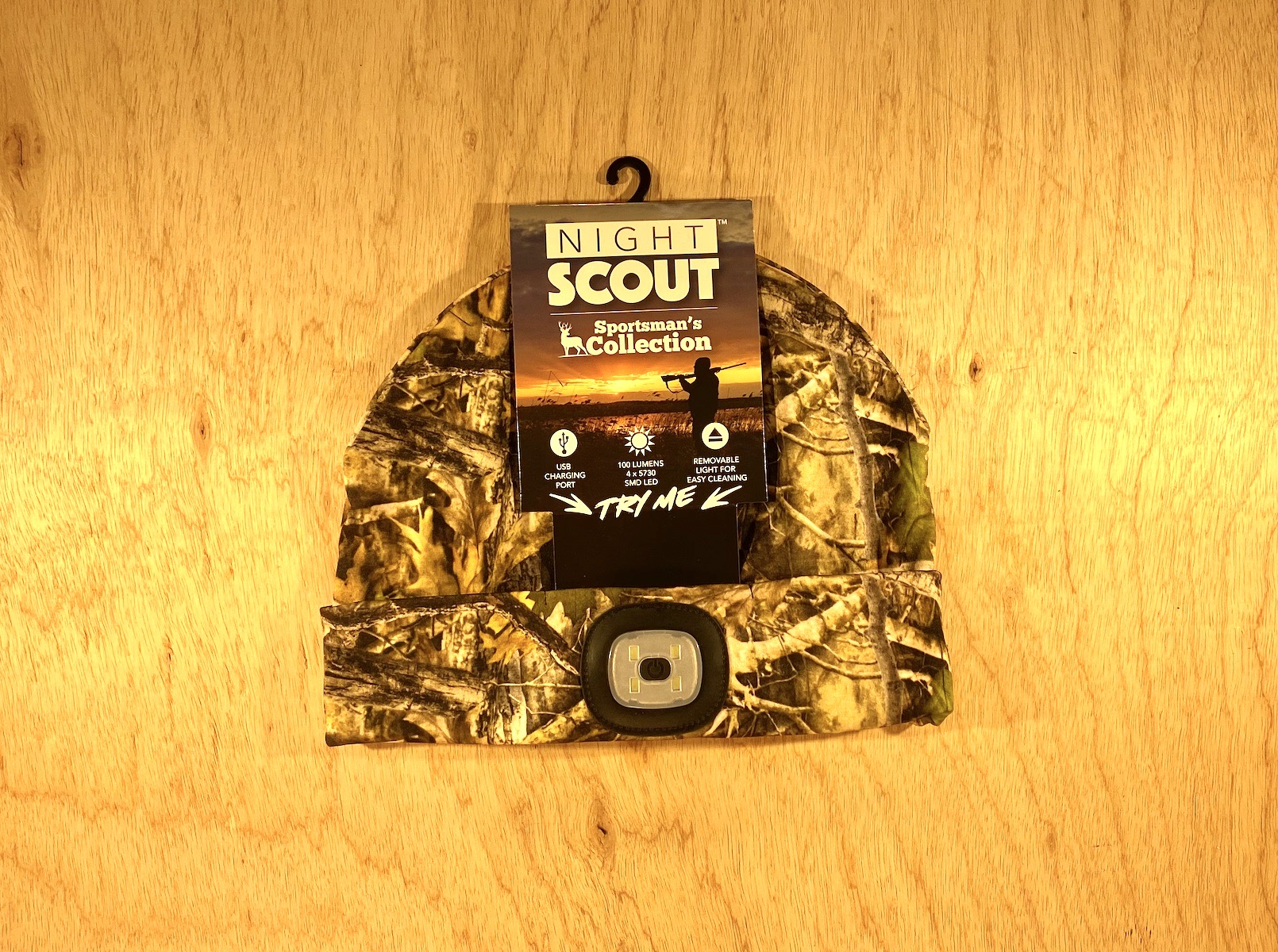 The Shelter Institute Night Scout Camo Hunting Headlamp Beanie is shown with its built-in LED light against a wooden background. The label shows the Night Scout Sportsmans Collection and a sunset silhouette.