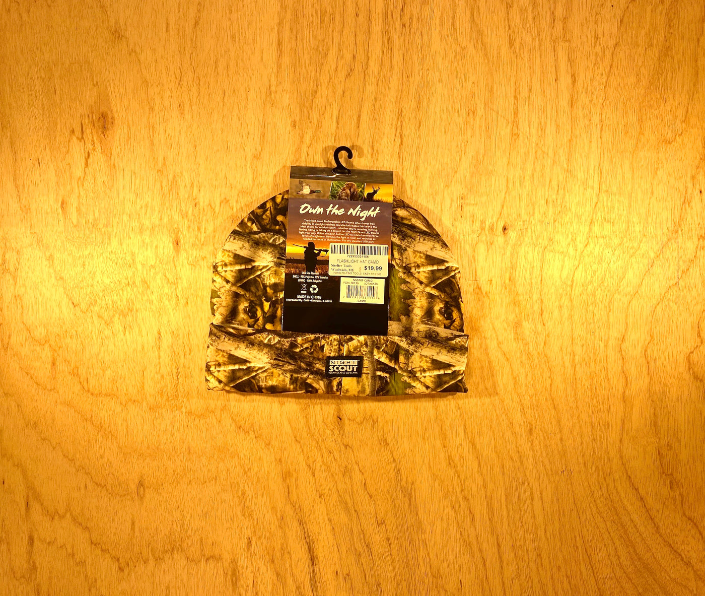 A Night Scout Camo Hunting Headlamp Beanie by Shelter Institute with a store tag and barcode, shown on light wood. The tag reads Out Here Night with an outdoor graphic—ideal for classic style lovers over LED beanies.