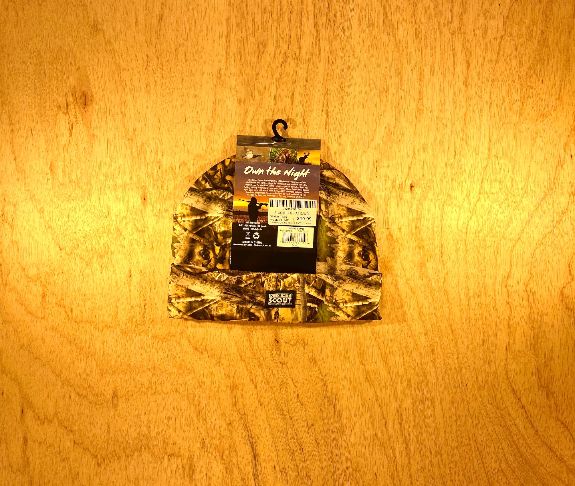 A Night Scout Camo Hunting Headlamp Beanie by Shelter Institute with a store tag and barcode, shown on light wood. The tag reads Out Here Night with an outdoor graphic—ideal for classic style lovers over LED beanies.