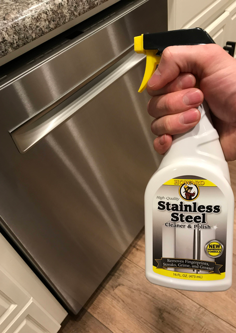 A hand holds a HOW Stainless Steel Cleaner spray bottle with a streak-free formula in front of a stainless steel dishwasher, ready to clean kitchen surfaces in a modern kitchen.