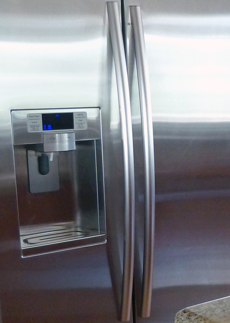 Close-up of a stainless steel refrigerator with two vertical handles and a water and ice dispenser. A granite countertop shines, thanks to HOW Stainless Steel Cleaner’s Streak-Free Formula for all kitchen surfaces.