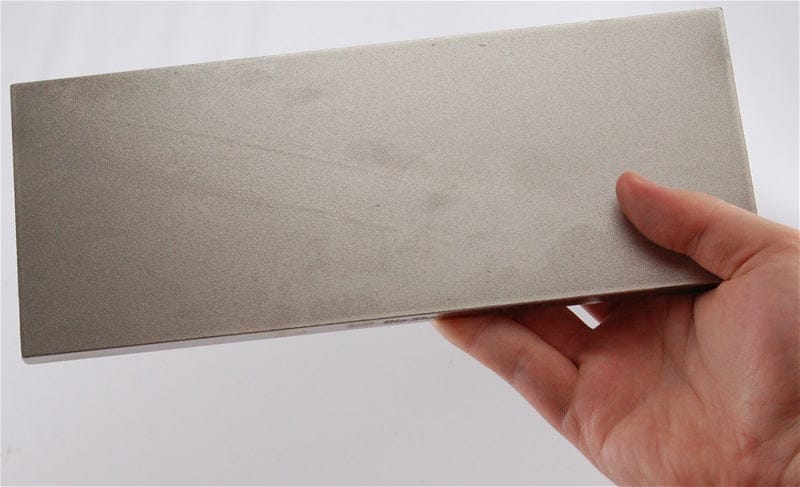 A hand holds a DMT Dia-Sharp Diamond Bench Stone 10-in, featuring a continuous diamond surface, against a white background.