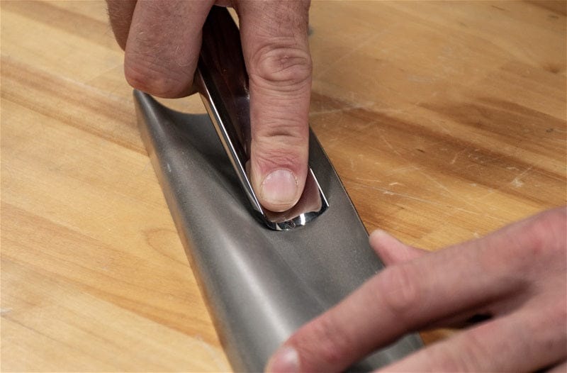 A person uses the DMT Diamond Wave sharpening tool to hone the edge of a large kitchen knife on a wooden surface.
