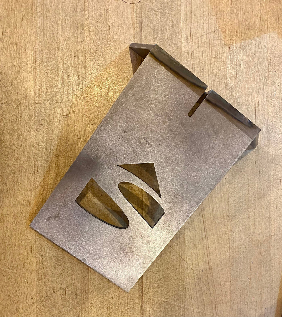 A Timber Frame Dovetail Layout Jig by Engineering Paige Trucks Restoration, featuring oval and triangle cutouts, rests on a wooden surface; its top edge is bent upward with two narrow slots near the bend.
