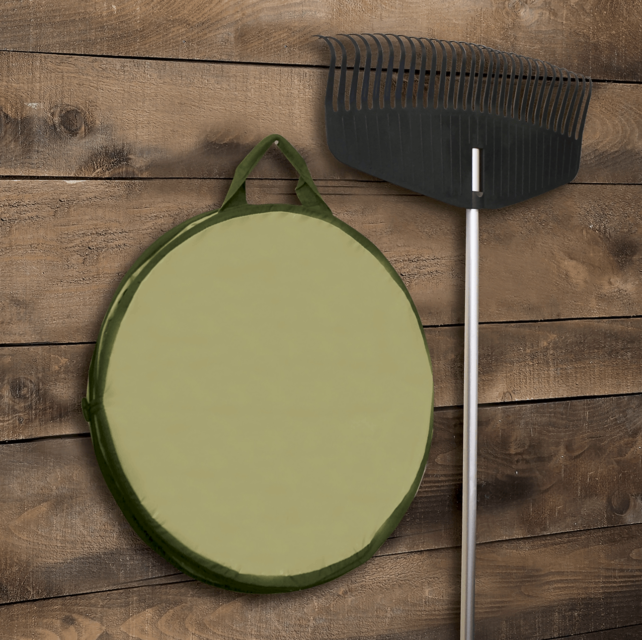 A rake with a black head and silver handle leans against a wooden wall, next to the tear-resistant Fiskars Kangaroo Gardening Container by FIS.