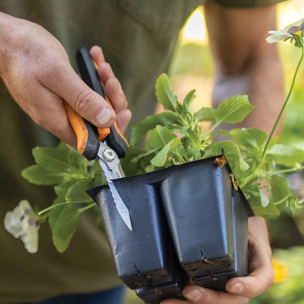 A person uses FIS Fiskars Multipurpose Garden Snips with stainless steel blades to cut a plastic plant container filled with green seedlings.