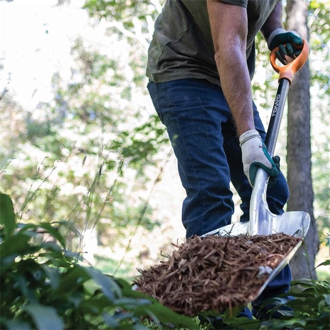 Wearing jeans and gloves, a person uses the FIS Scoop Shovel with an aluminum head to move mulch in the garden.