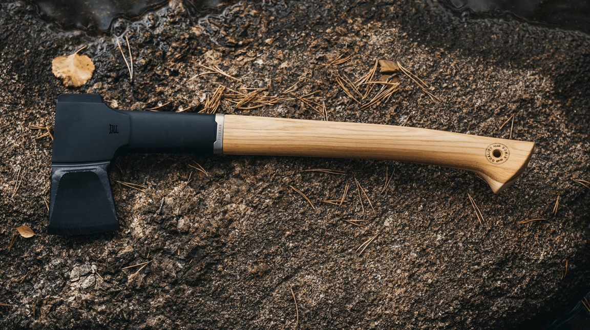 The FIS Fiskars Norden N12 Splitting Axe, featuring a black metal head and hickory wood handle, rests on a textured rock surface surrounded by scattered pine needles.