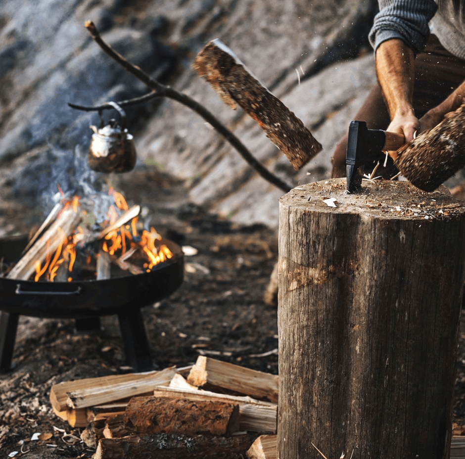 A person uses a Fiskars Norden N12 Splitting Axe from FIS, featuring a hickory handle, to chop wood near a campfire with a kettle hanging over the flames.