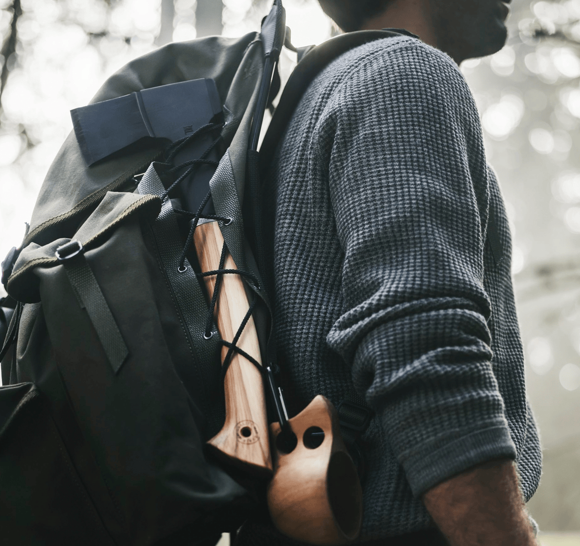Wearing a gray sweater, the person strides through the forest with a backpack carrying a FIS Fiskars Norden N7 Hatchet expertly secured on the side, showcasing fine axe craftsmanship.