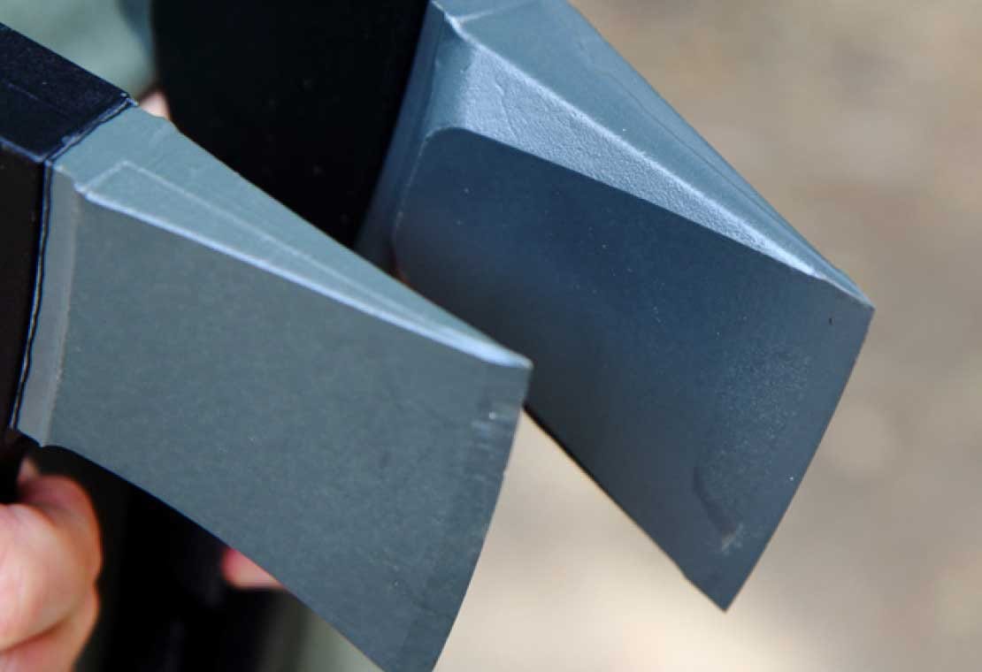 Close-up of the metal blades on two FIS Fiskars Splitting Axe 23-in Handle axes, side by side with ultra-sharp edges and matte grey finish; a hand is partially visible gripping one axe’s handle.