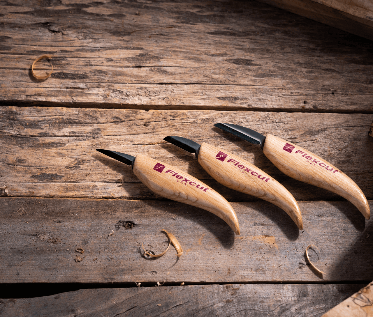 The 3-Knife Starter Set from FLE includes three wood carving knives, each with an ergonomic wooden handle, elegantly displayed on a rustic surface surrounded by wood shavings. Made with edge-holding steel, these knives provide precision and durability, making them ideal for both beginners and experienced artisans.