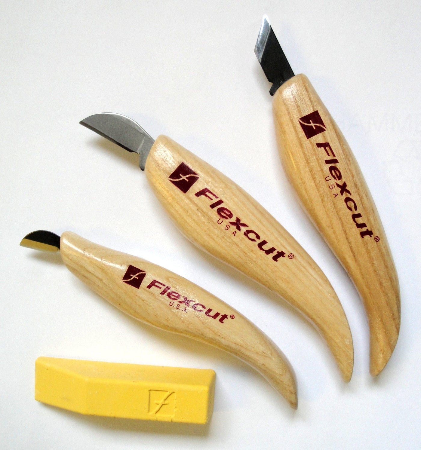 Three FLE Chip Carving Set knives with curved wood handles and a small yellow sharpening strop are arranged on a white background.