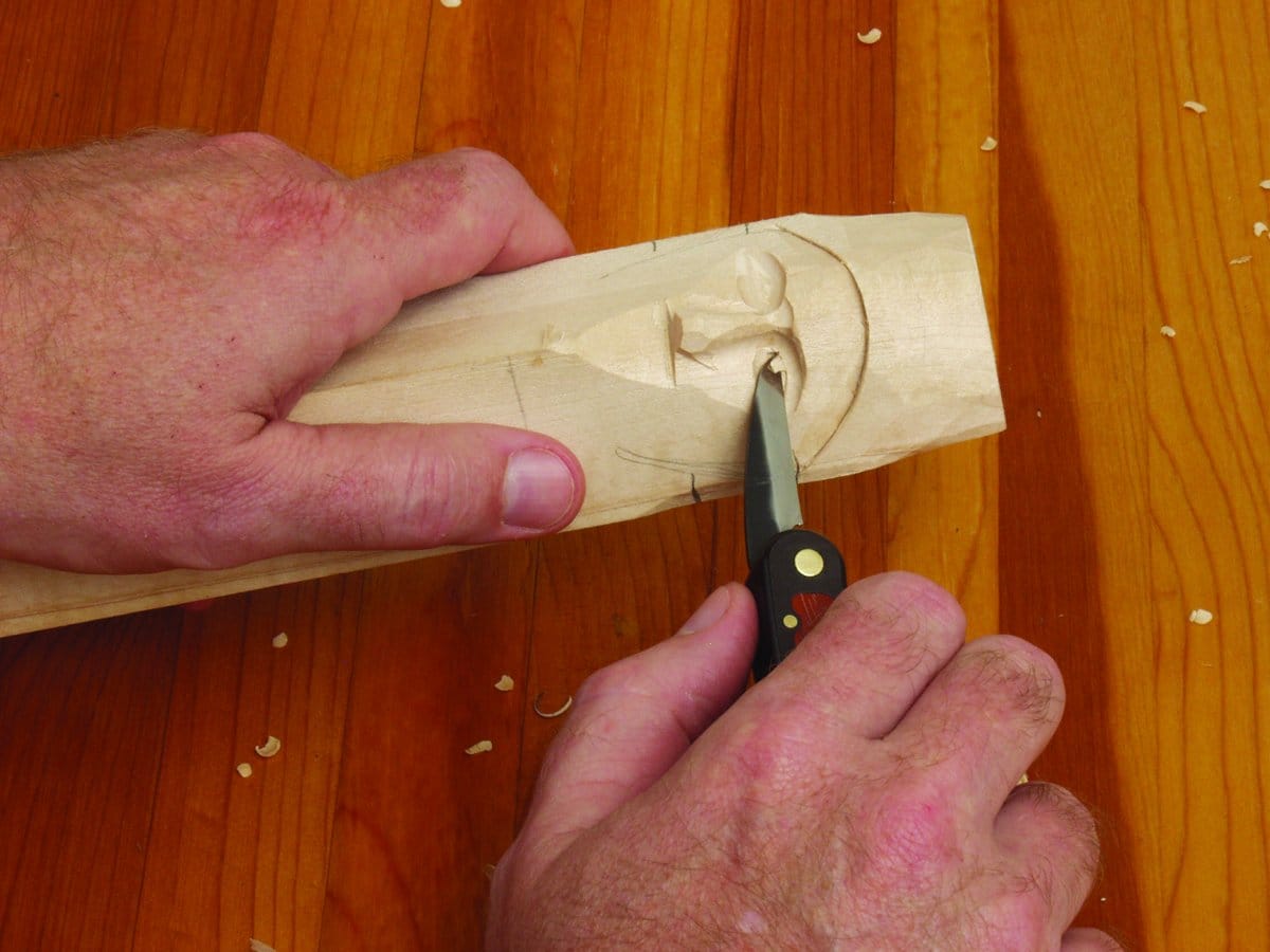 Using the FLE Carvin Jack, a person whittles a human face into light-colored wood on a wooden surface, with wood shavings scattered around their hands and the carving.