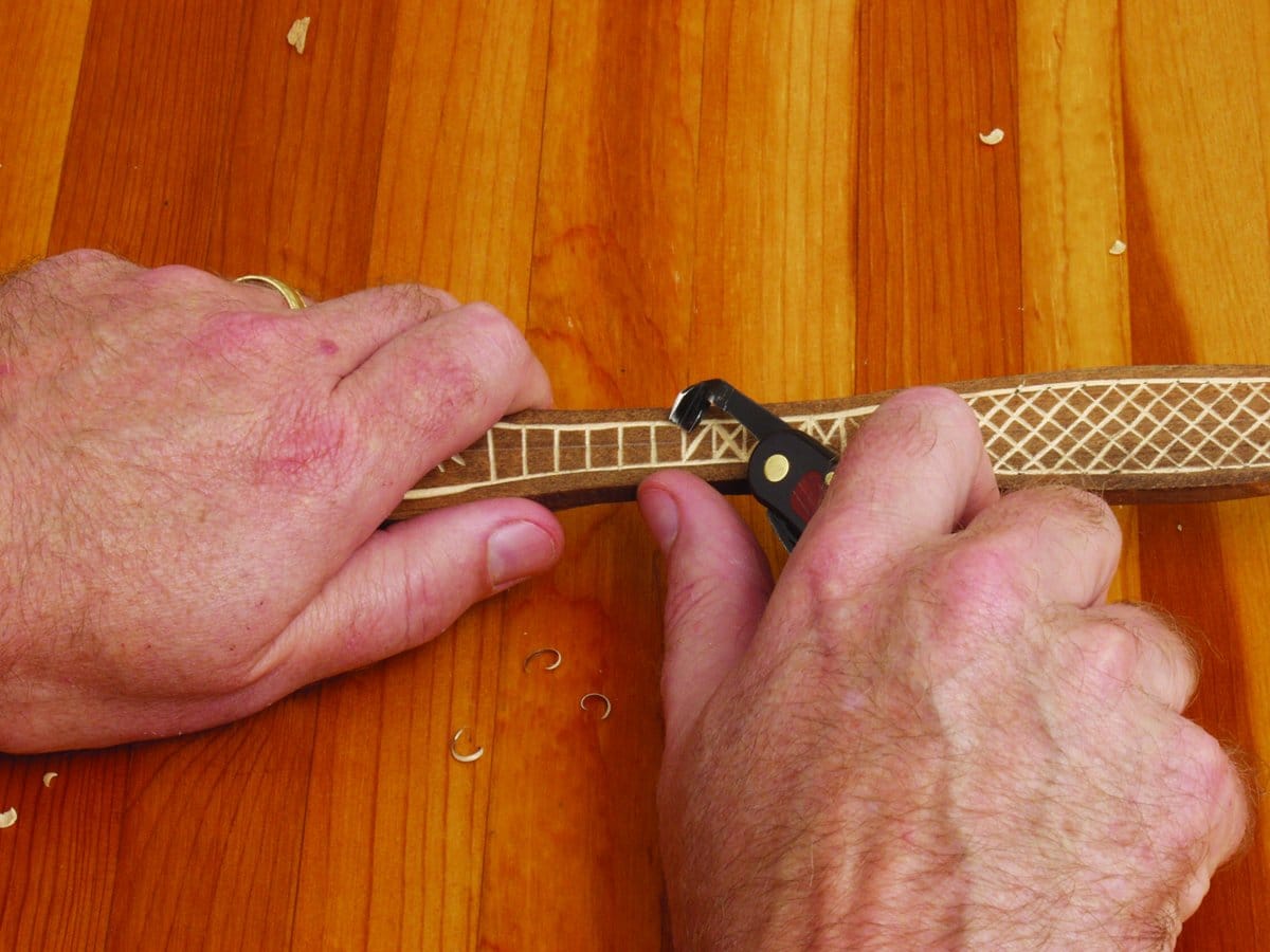Close-up of hands using the FLE Carvin Jack multi-tool to carve patterns into a wooden stick on a wooden surface, with small wood shavings scattered around.