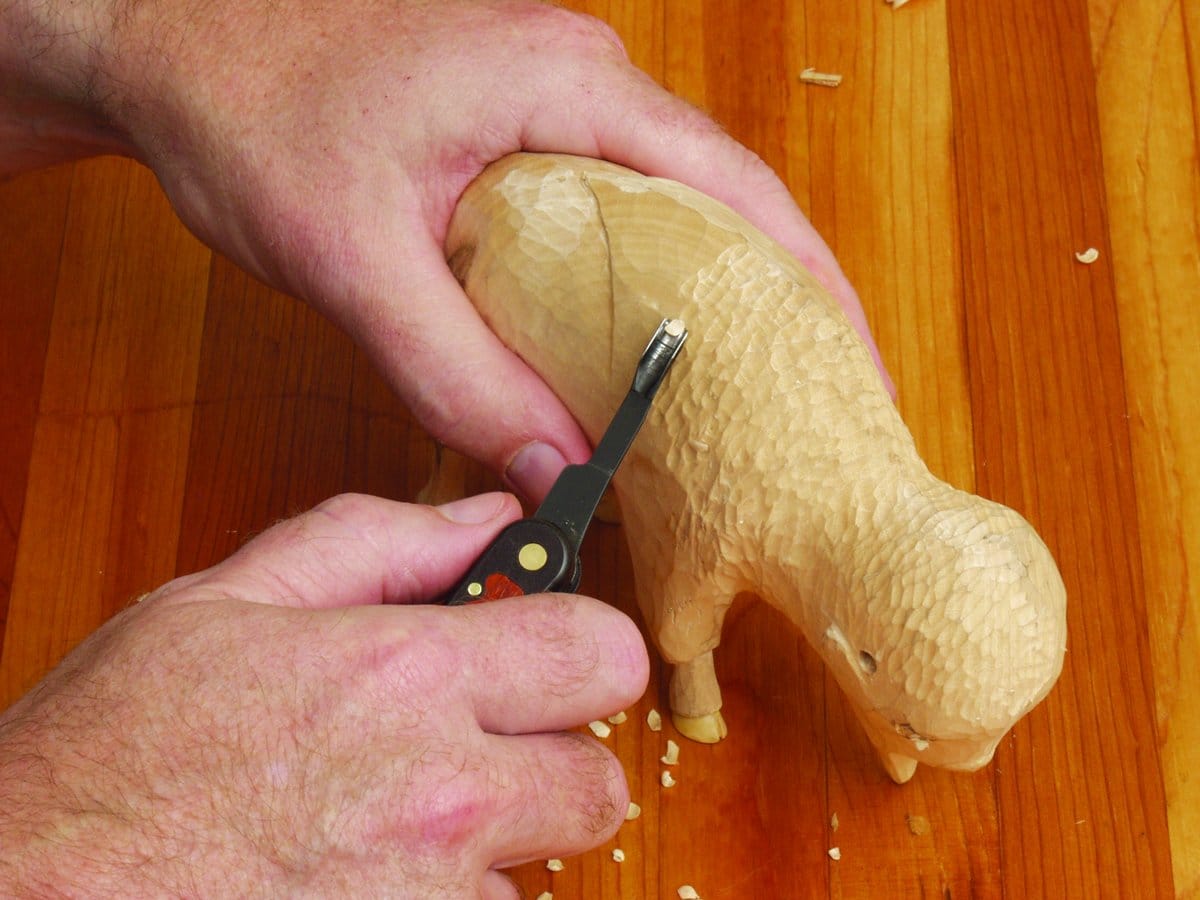 Using the FLE Carvin Jack, someone carves fine details into a wooden sheep figure on a wooden surface, surrounded by wood shavings.