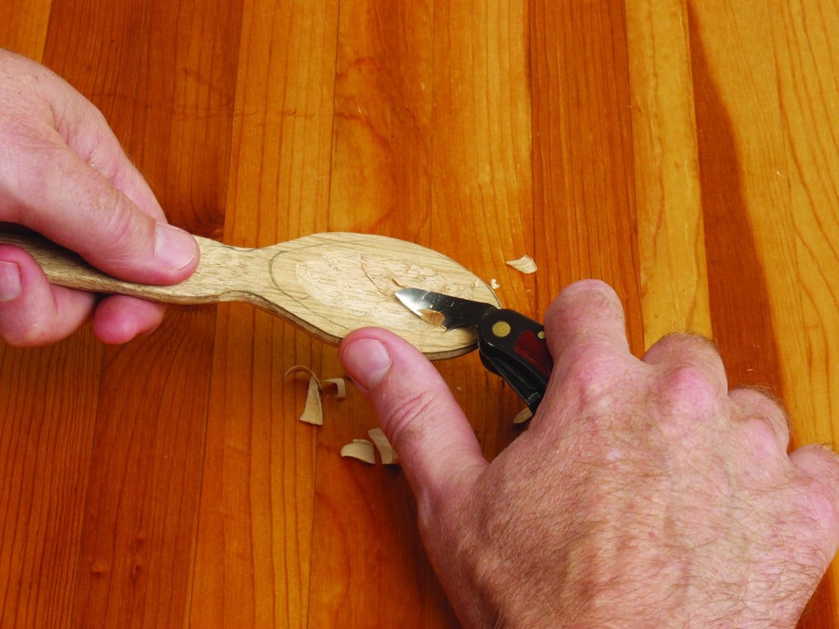 Using the FLE Carvin Jack, a person carves the bowl of a wooden spoon on a polished wood surface, surrounded by scattered wood shavings.