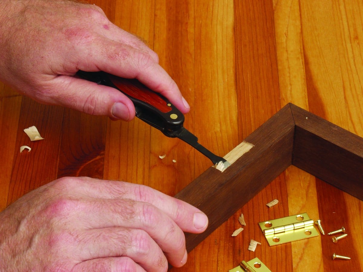 Using the FLE Carvin Jack folding pocket knife, a person carves wood from a frame’s corner while brass hinges and screws rest on the wooden surface nearby.