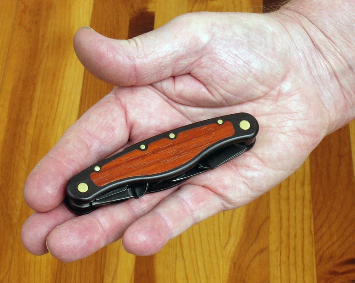A person’s hand holding a closed FLE Carvin Jack pocket knife with a wooden handle and brass rivets over a wooden surface—an essential woodcarving multi-tool.